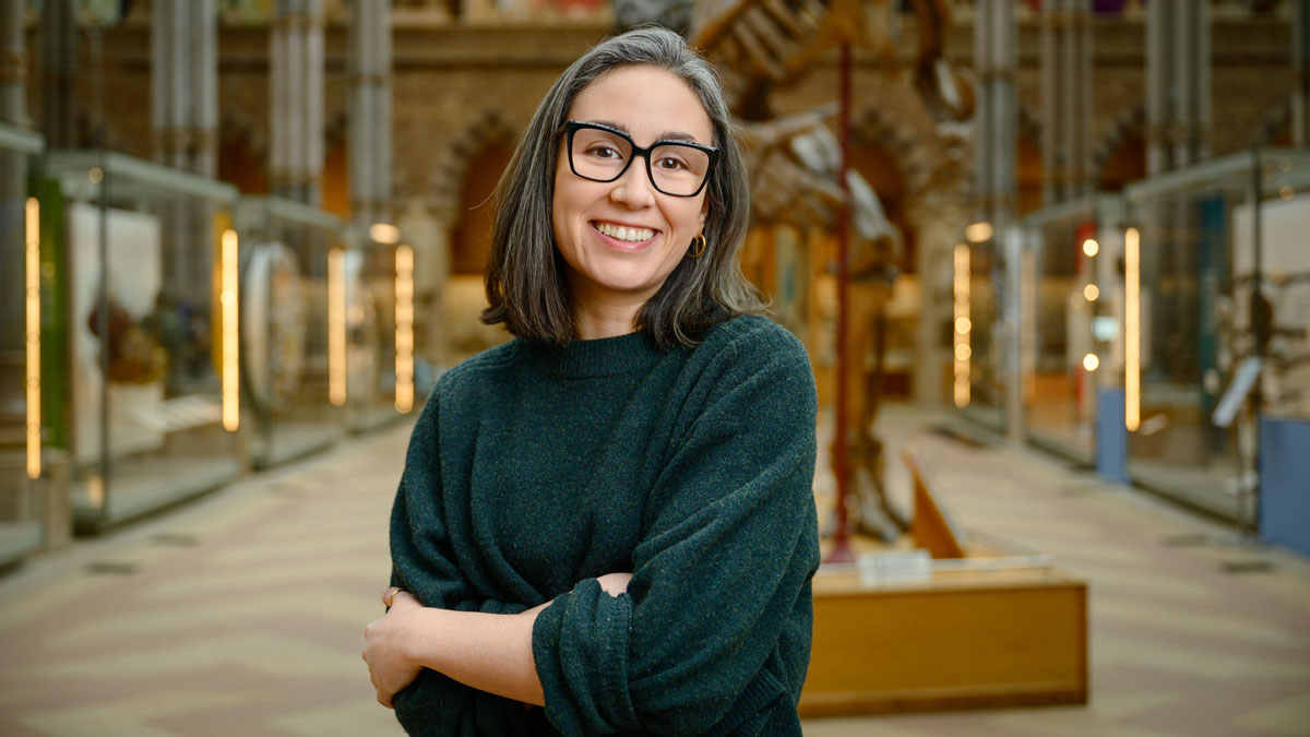 Lucia Perez Diaz, wearing a green sweater, stands in front of a dinosaur skeleton in a natural history museum.