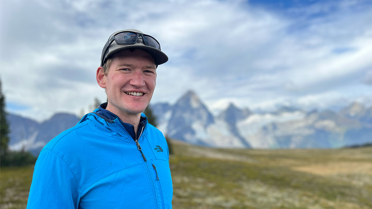 A man in a hat and blue jacket stands, smiling, in front of a mountain range.