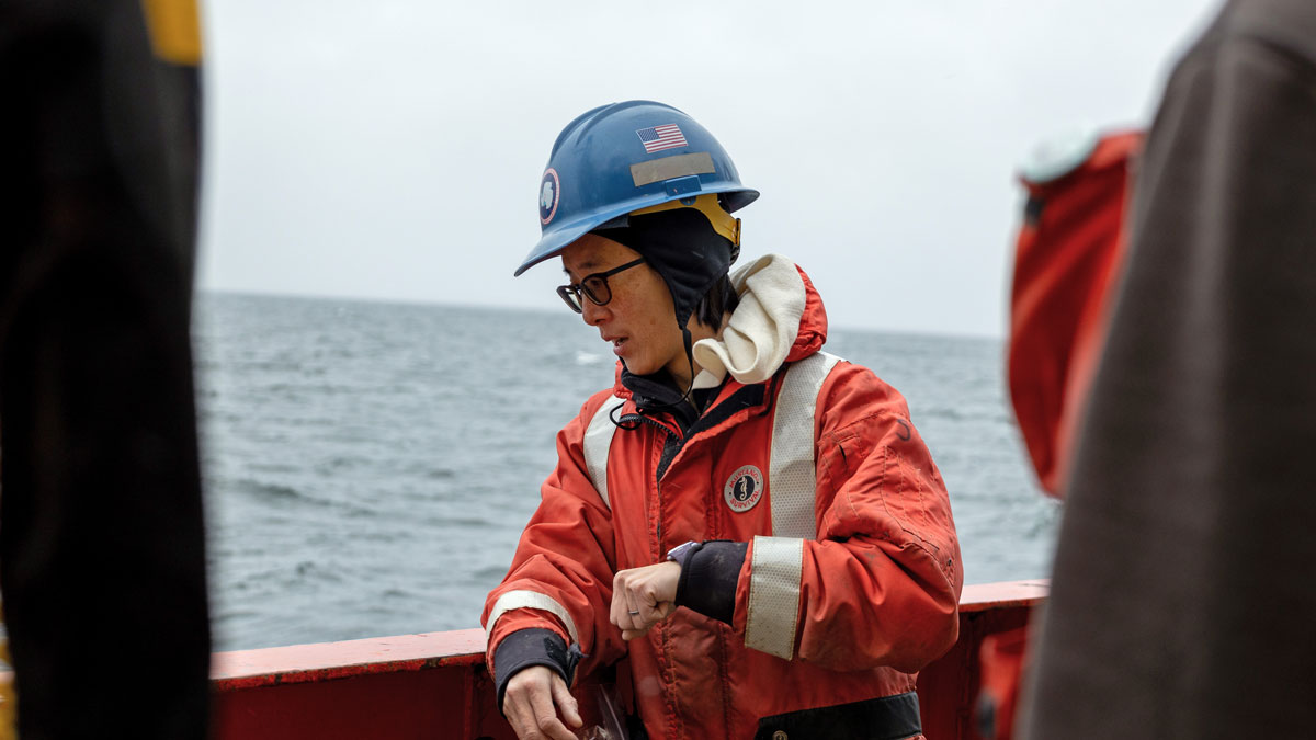A woman in a blue helmet and orange jacket looks over the edge of a ship.