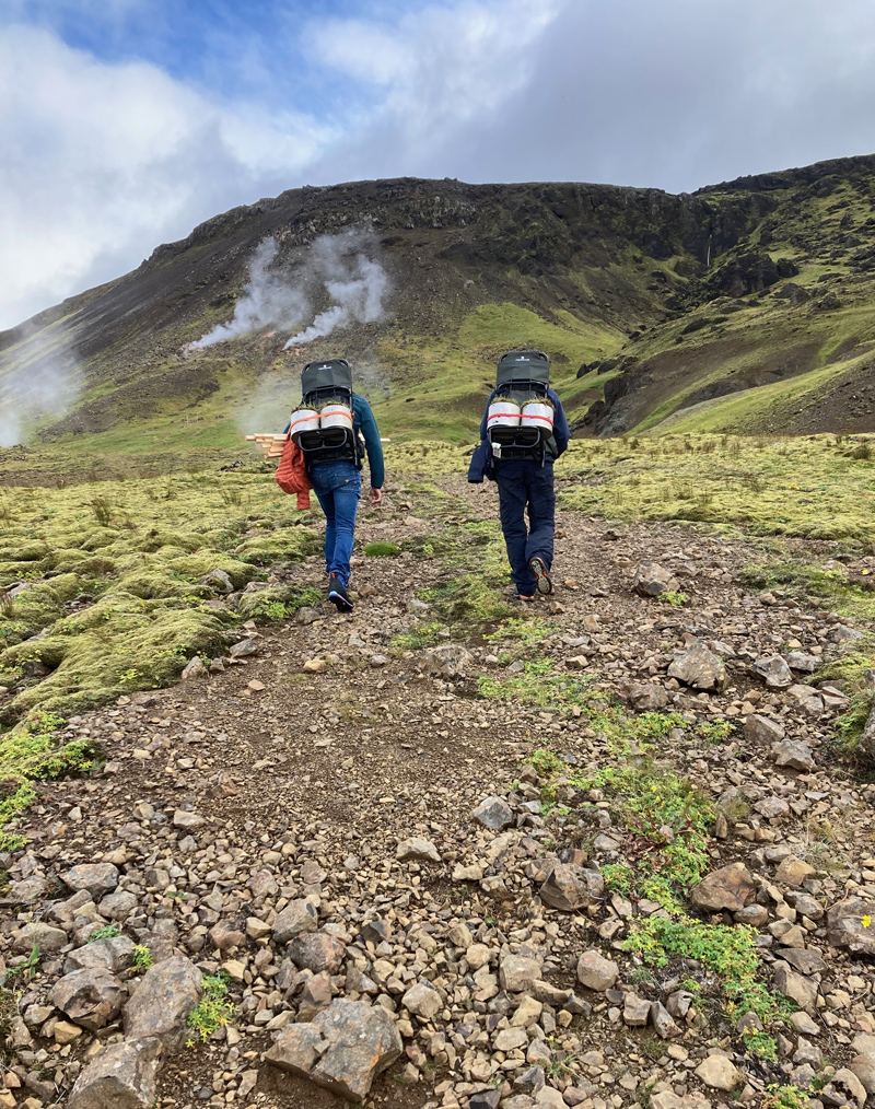 Two people walk up a patch of soil on a green hillside with small tanks on their backs.
