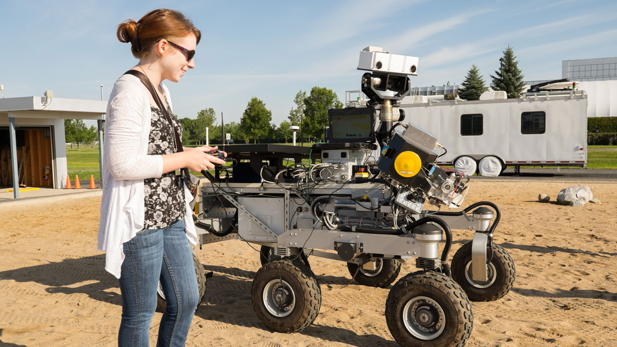 A person wearing sunglasses uses a remote control to drive a chest-height, six-wheeled rover on dirt.