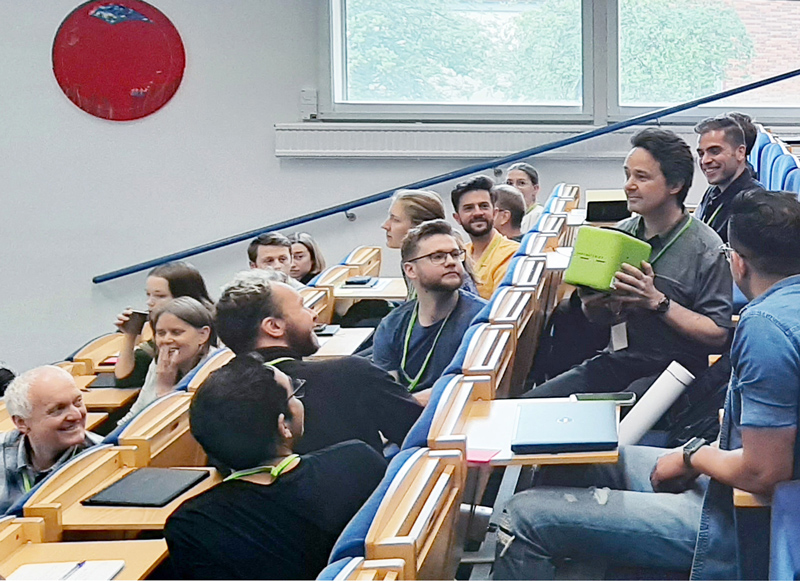 People sit in several rows of amphitheater-style seating in a classroom. One holds a green, cube-shaped microphone.