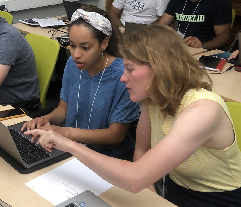 Two people sitting at a desk look at a laptop monitor.