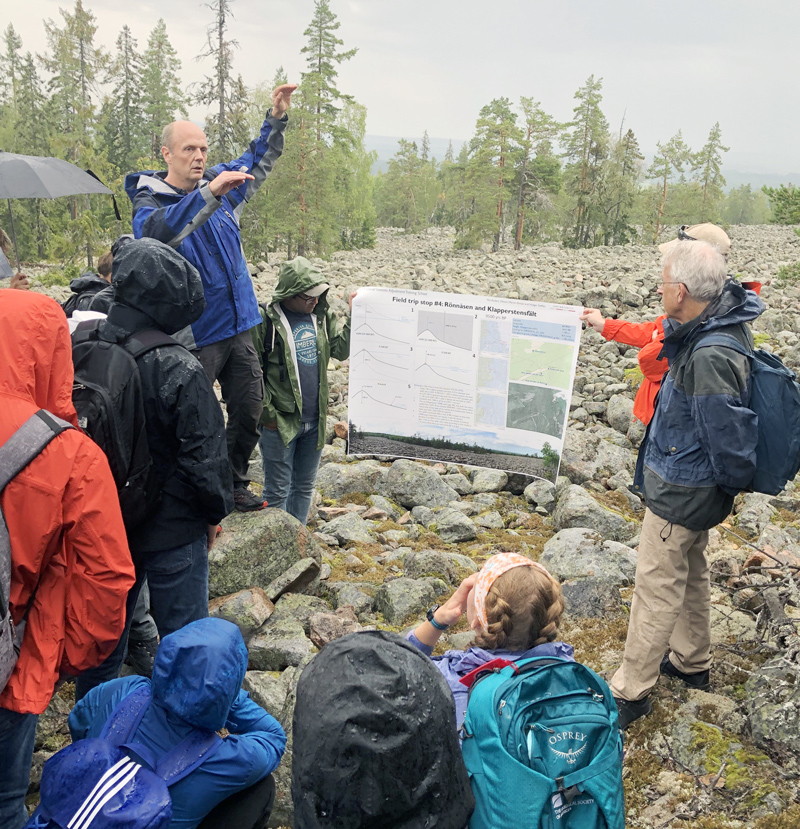 An instructor and a group of students stand in a rocky clearing amid patchy tree stands discussing the local geology while two people hold up a poster with information about the landscape.