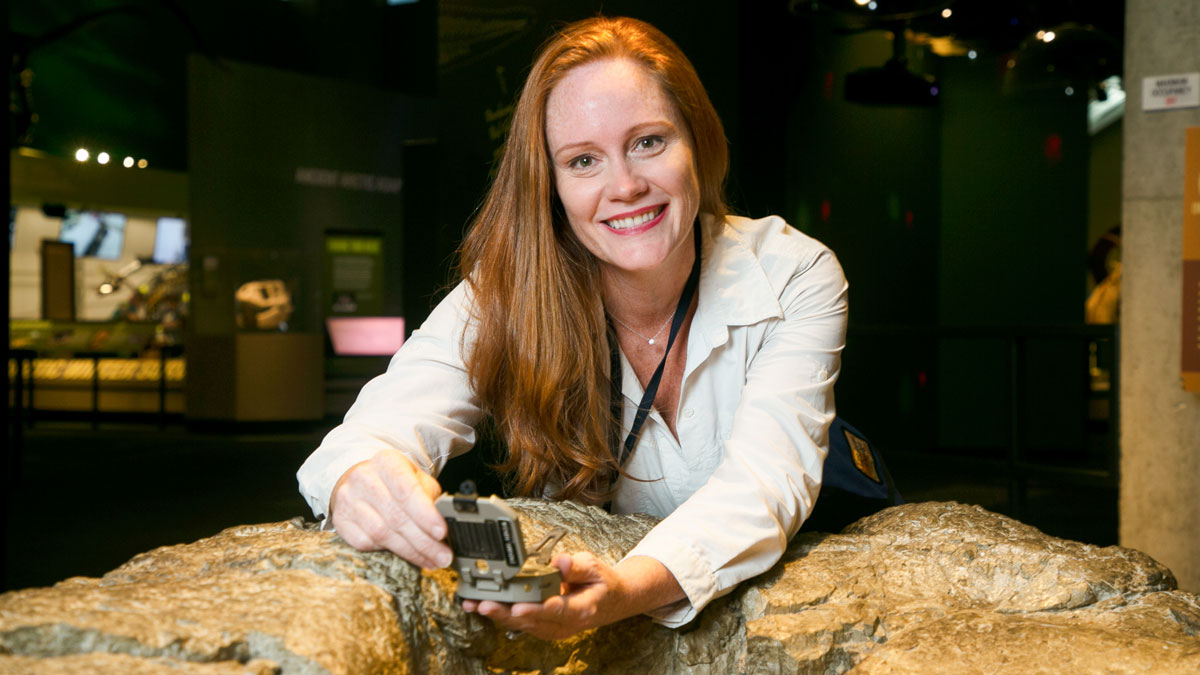 A red-haired woman holding a geologist’s compass and looking at the camera leans over a rock inside a building.