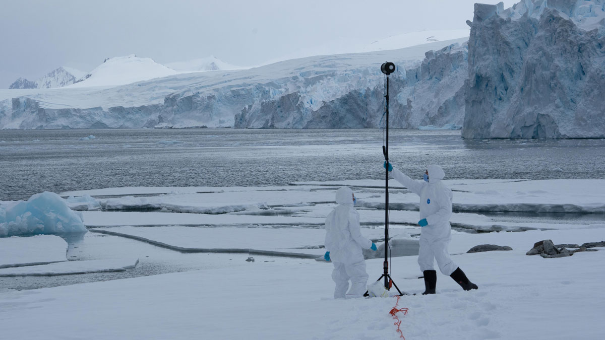 Two people stand in a snowy landscape near icy water and glaciers, holding up a black pole that is about twice as tall as them.