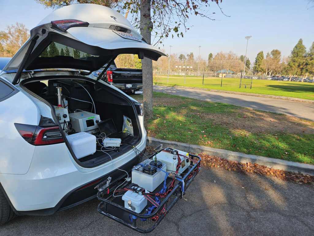 A white hatchback vehicle has a metal frame attached to the back of it. The frame and the car both hold boxy instruments connected to one another with wires.