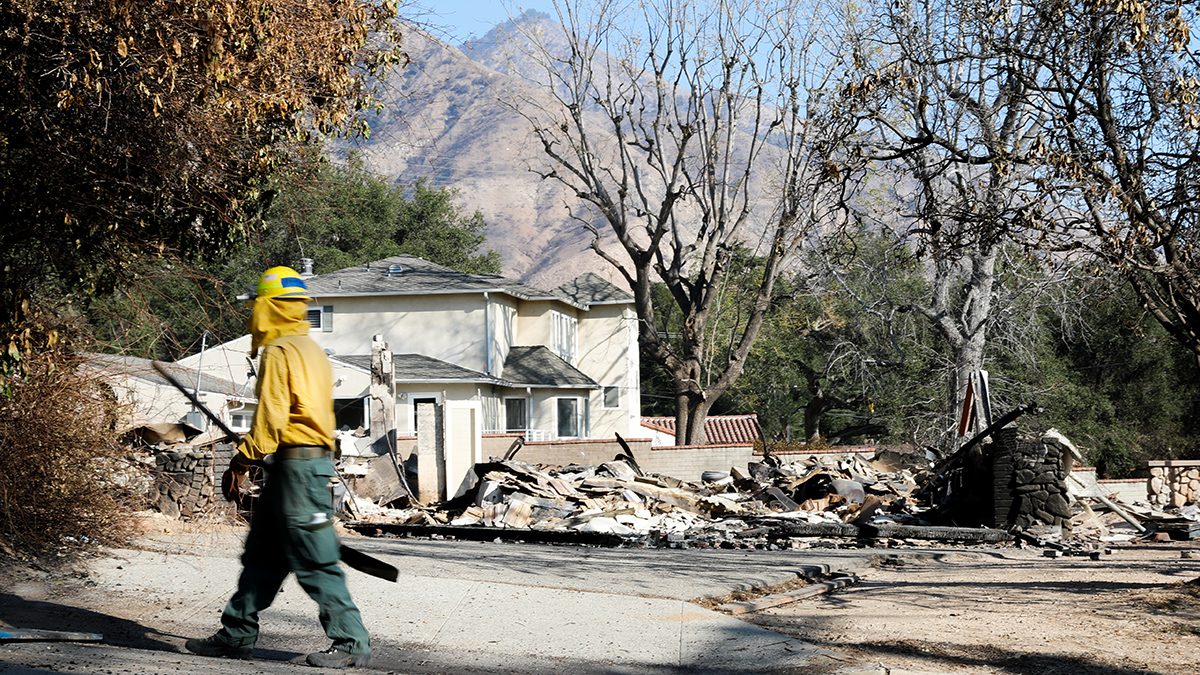 A person in yellow and green protective gear walks past a semi burned house with a mountain backdrop.
