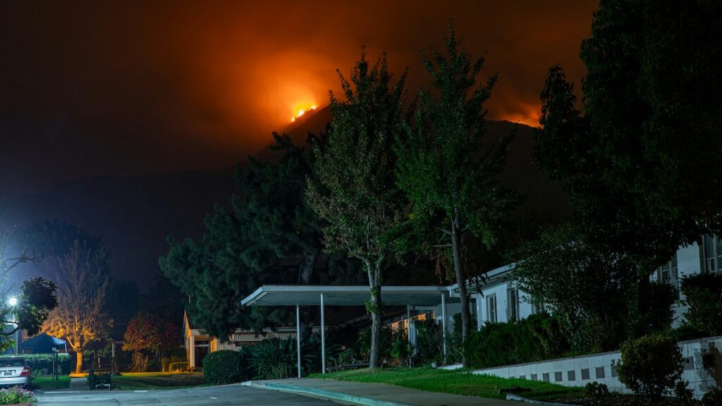 A street and a building in the foreground, with a fire burning atop a mountain in the background.