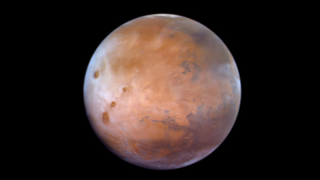 In this bird’s-eye satellite view, light brown peaks and valleys appear beneath a thin, grayish-white haze of foggy cloud cover that varies in translucence.