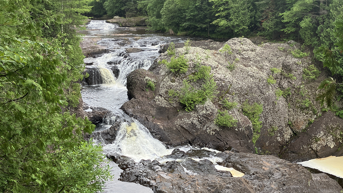 A waterfall runs over red and gray rocks. Evergreen trees line the fall’s edges.