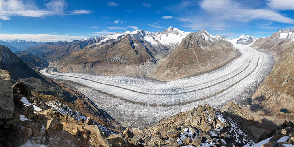 Panoramic view of a long glacier flowing through a valley among tall mountain peaks under blue sky.