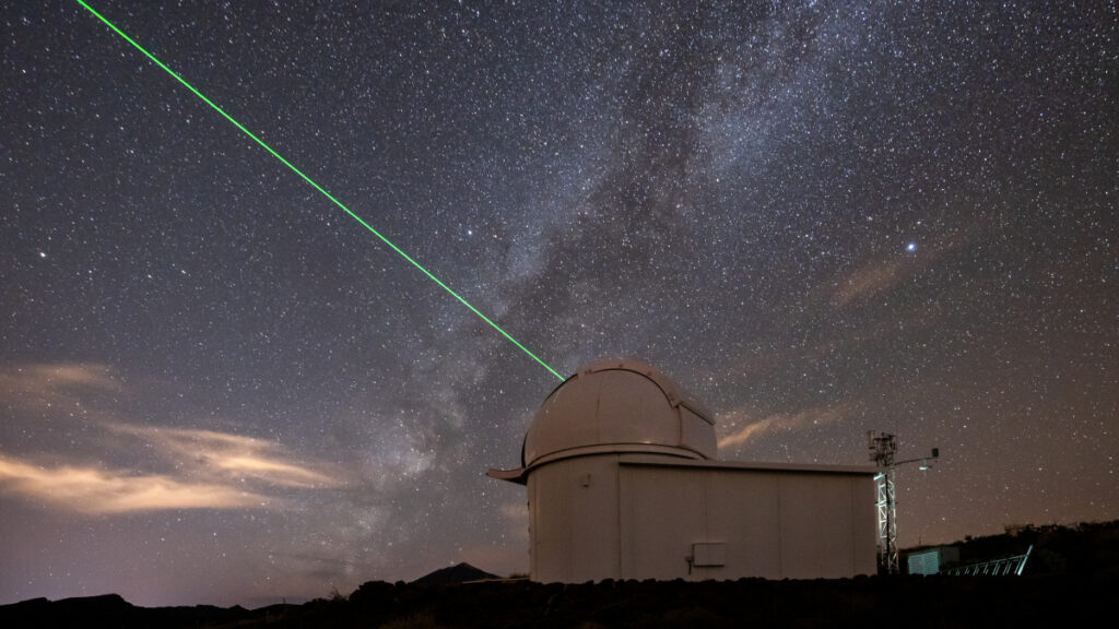 A telescope dome emits a green laser beam into the starry night sky.