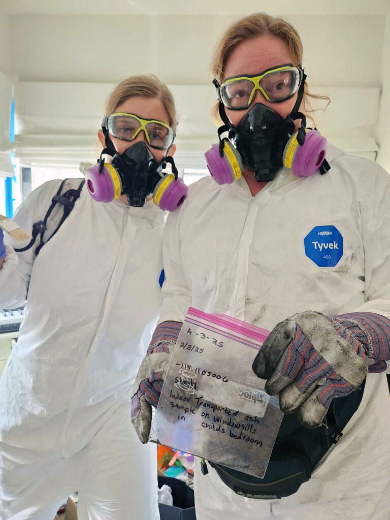 Two women wear Tyvek suits, goggles, gloves, and masks to filter out fine particles. One is holding a Ziploc bag that reads, “Indoor transported ash samples on windowsills in child’s bedroom. 2/3/25.”