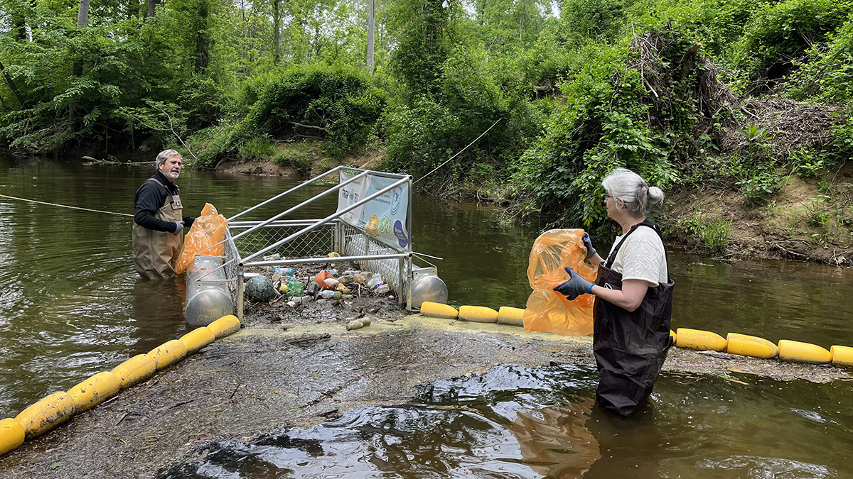 Two people wearing waders stand in a river holding orange trash bags. Between them is a cage flanked by two lines of buoys, which have funneled trash into the cage.