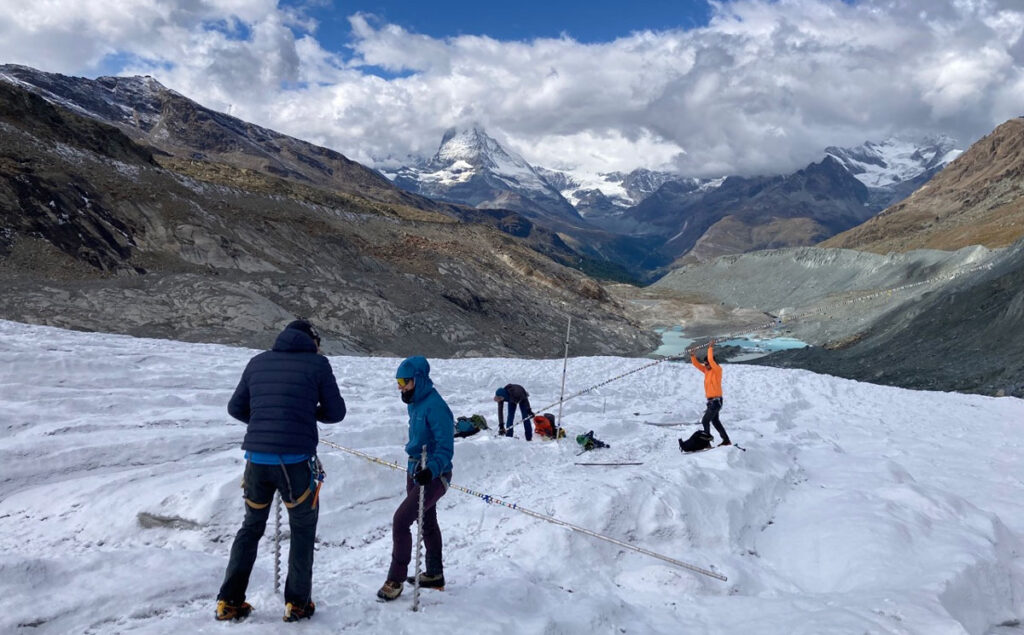 Four researchers stand atop a glacier set among tall mountain peaks, preparing to install long poles called ablation stakes.