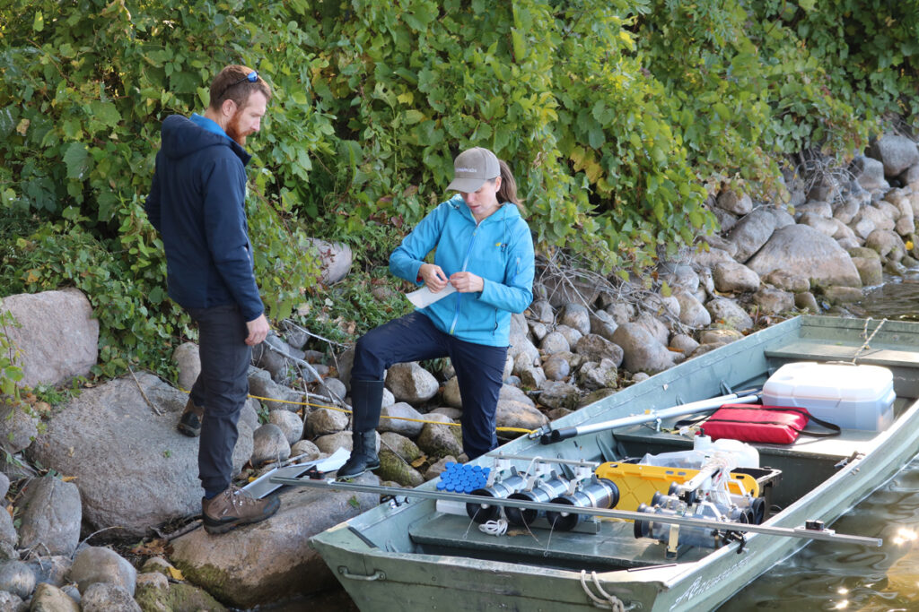 A man and a woman on a lakeshore stand by a rowboat loaded with water sampling equipment.