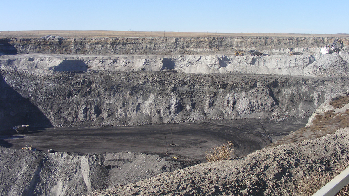 A large surface mine shows a deep hole with multiple trucks and excavation equipment that are dwarfed compared to the size of the mine pit