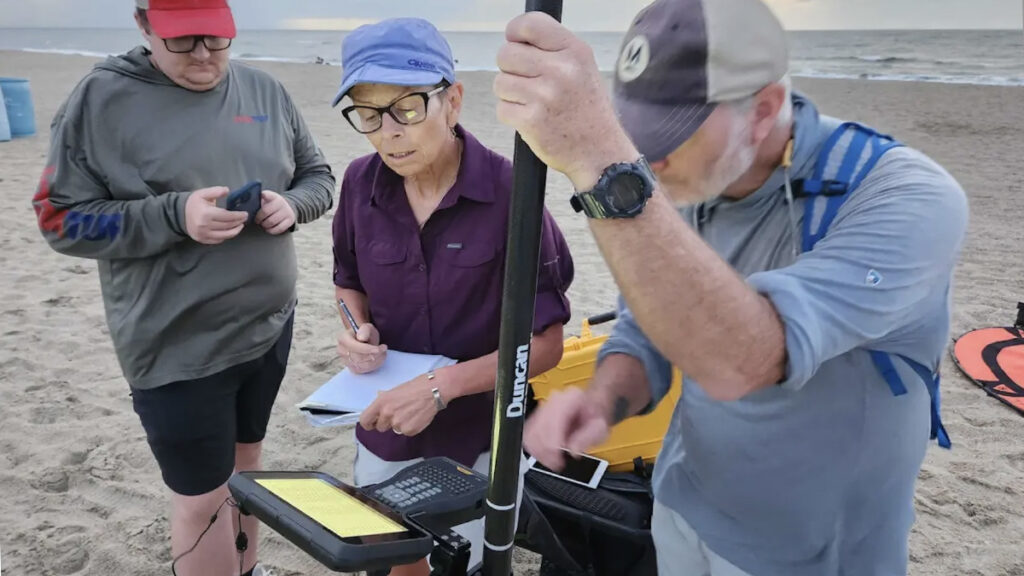 Three researchers stand on a cloudy beach, examining a set of lidar equipment.