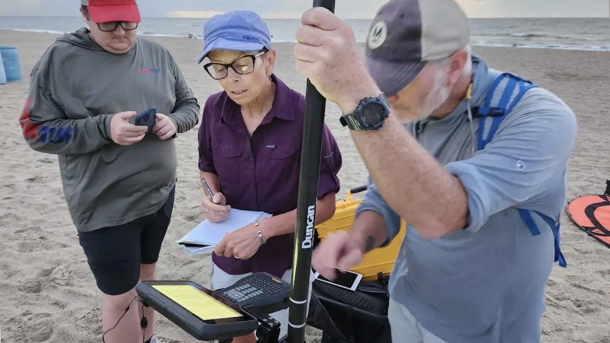 Three researchers stand on a cloudy beach, examining a set of lidar equipment.