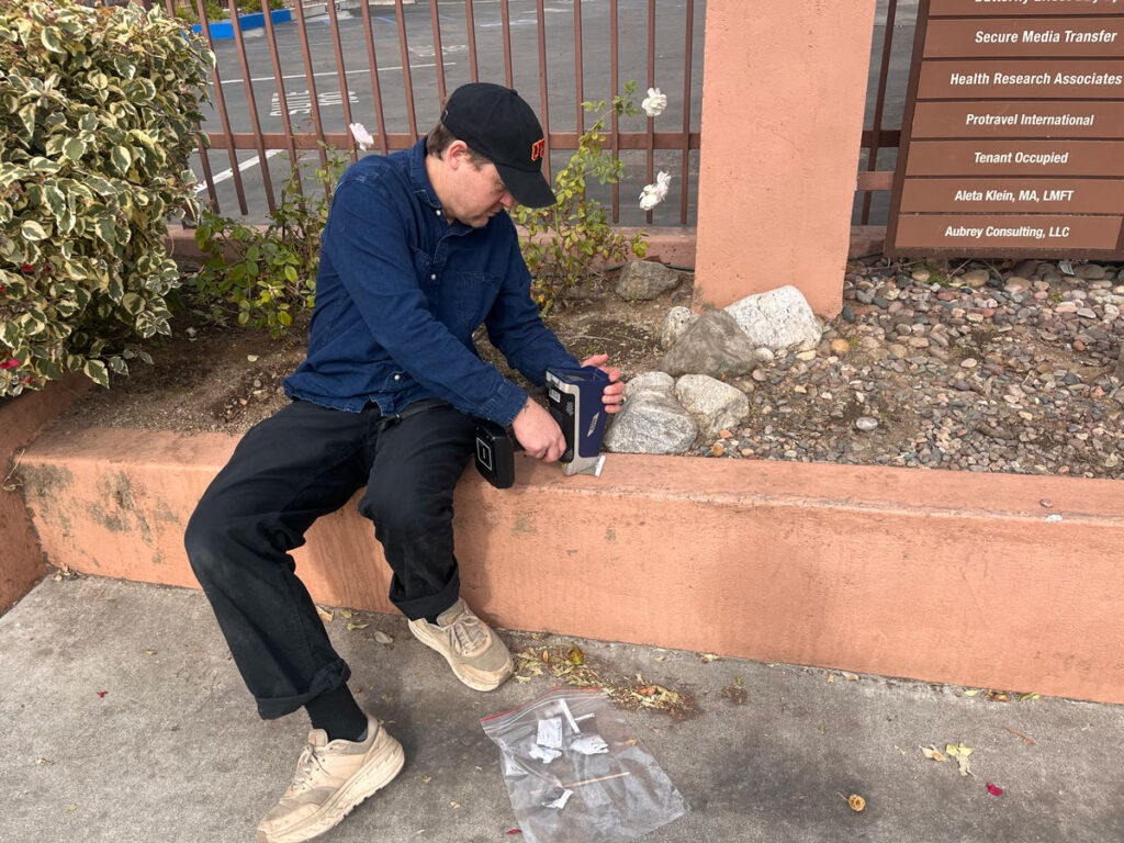 A person sits outside on a low wall and holds a handheld scanner to a small bag of gray dust.
