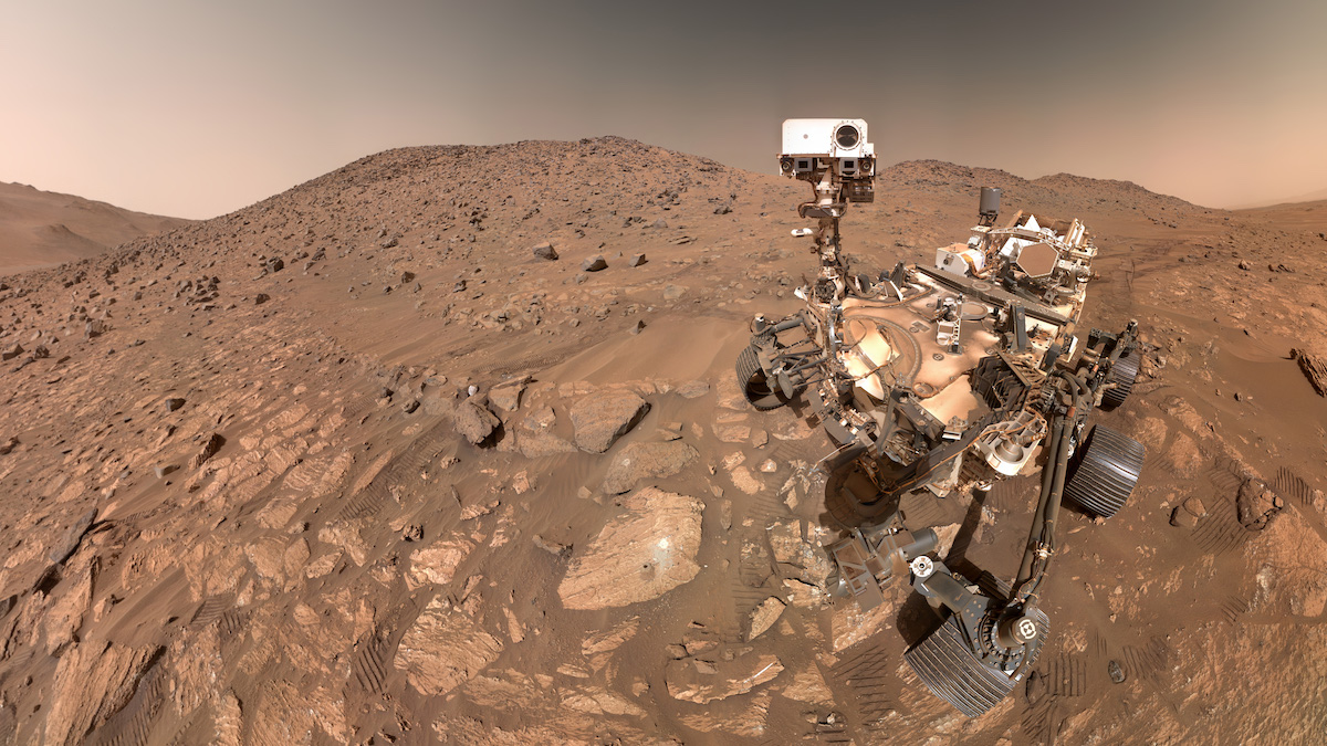 The Perseverance Mars rover, a robot, pictured in front of a red landscape on Mars.
