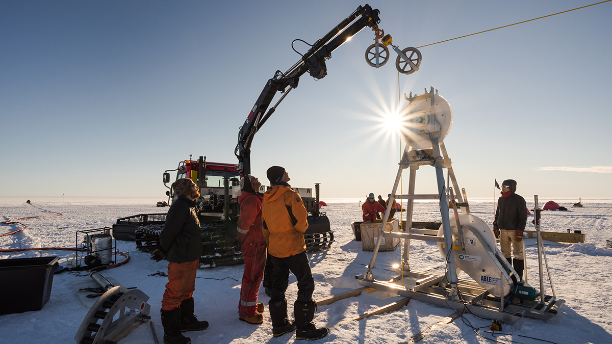 Four warmly dressed scientists stand around a large metal rig taller than them that is drilling a hole into the ice they are standing on. More scientists stand in the background, and a crane is stretched over the metal rig, lowering a cable into the hole.