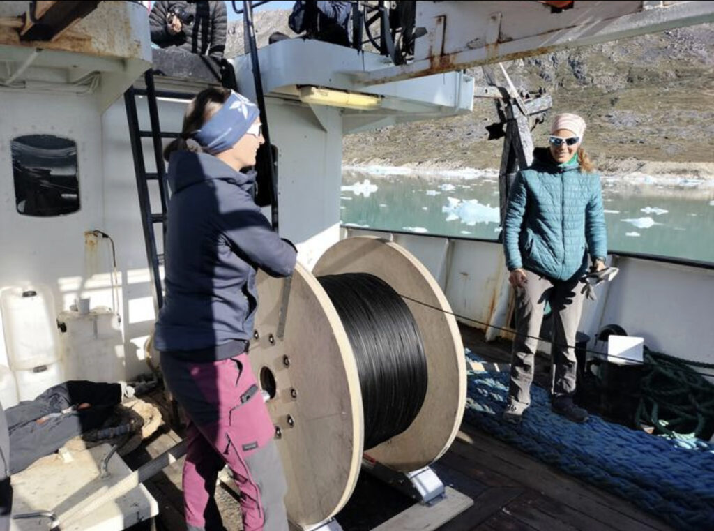 Two women deploy a long fiber-optic cable from the bow of a ship.