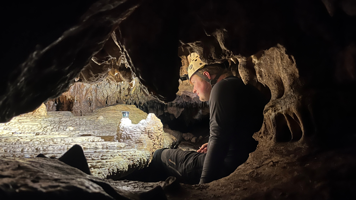 A researcher in a hard hat sits in a backlit cave with a small metal instrument.