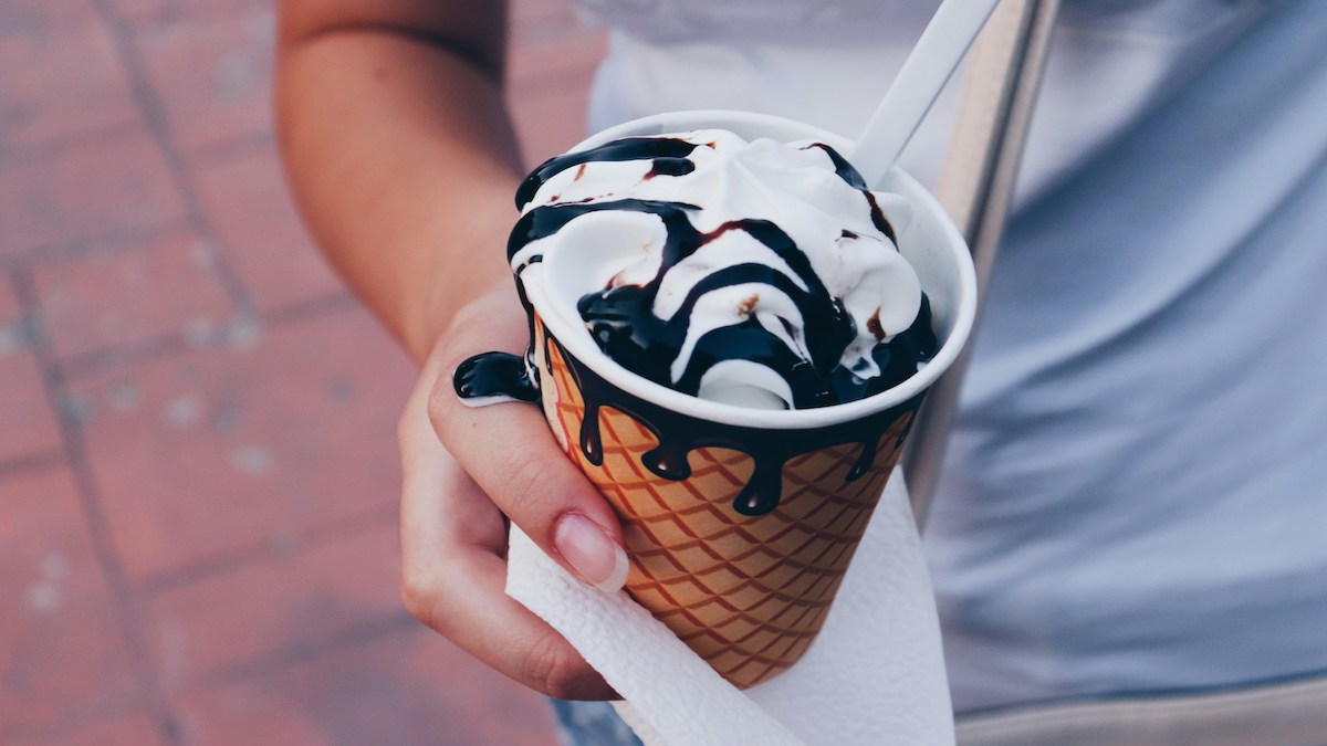 A person’s hand holds a cup with ice cream near a sidewalk in a city.