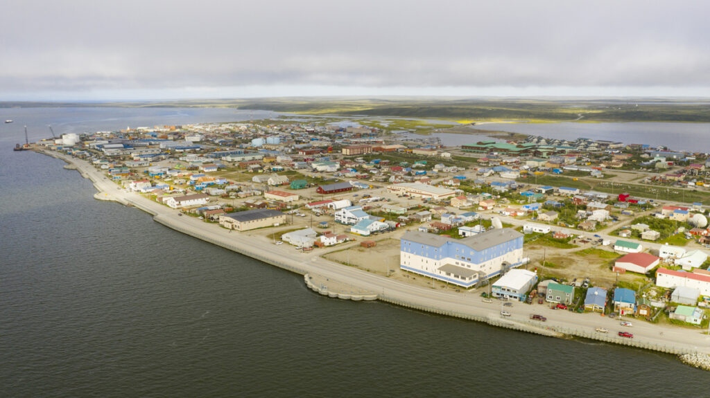 Aerial view of a small coastal town in Alaska, with low-lying buildings and homes and a roadway following the shoreline