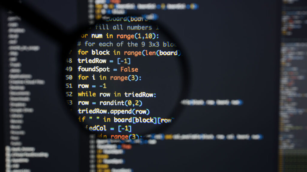 A magnifying glass is held up in front of a computer screen displaying lines of code.