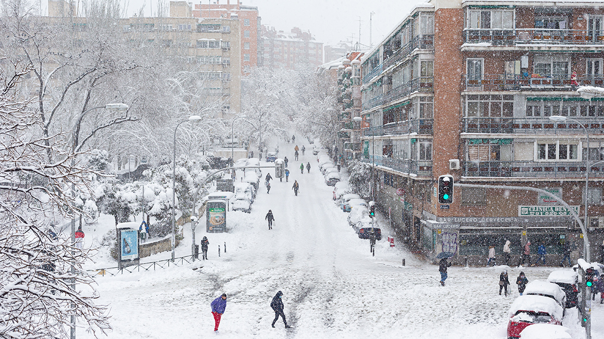 People walk down the middle of snow-covered streets among trees and several-story buildings while snow continues falling.