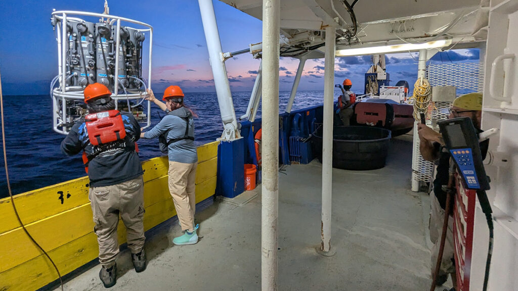 Two people on the deck of a boat around sunset reach for an instrument—almost as tall as they are—being raised up over the side of the ship from underwater.
