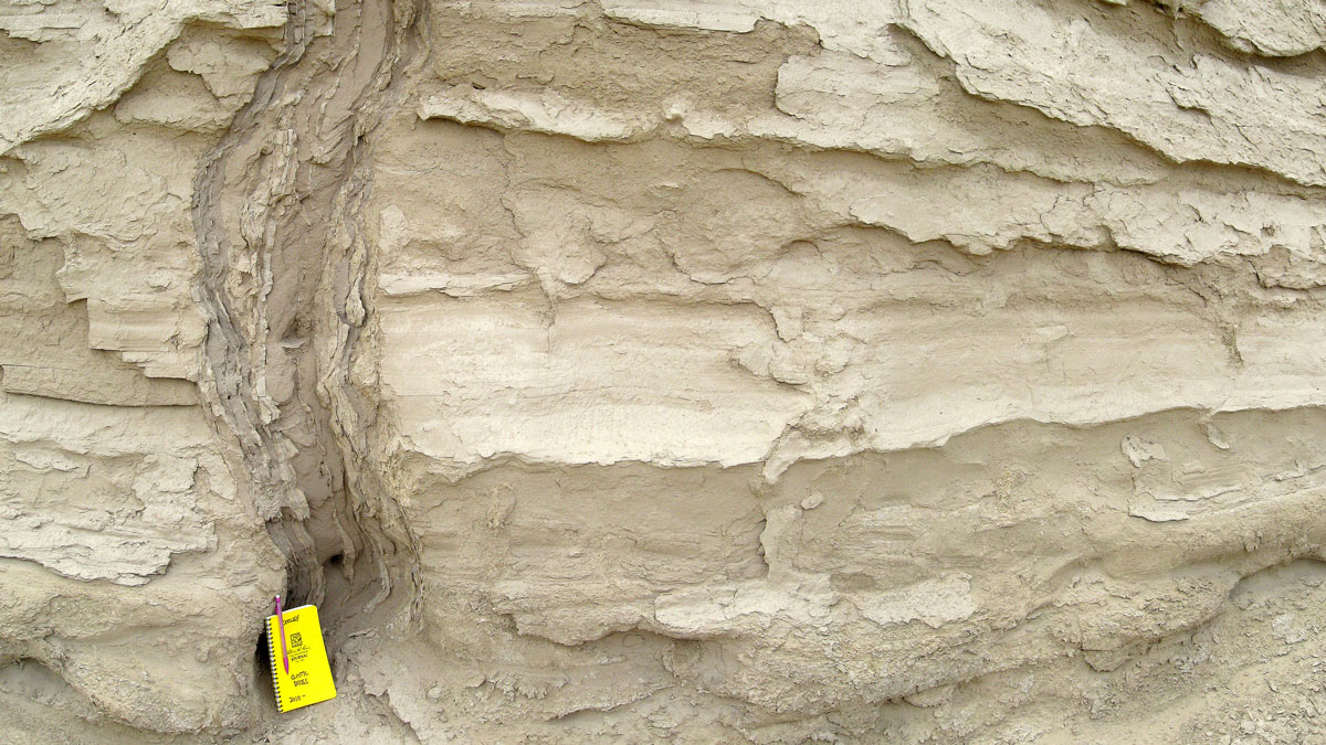 Layers of beige-colored rock with a vertical band of darker-colored rock. A yellow notebook appears at the bottom of the image.