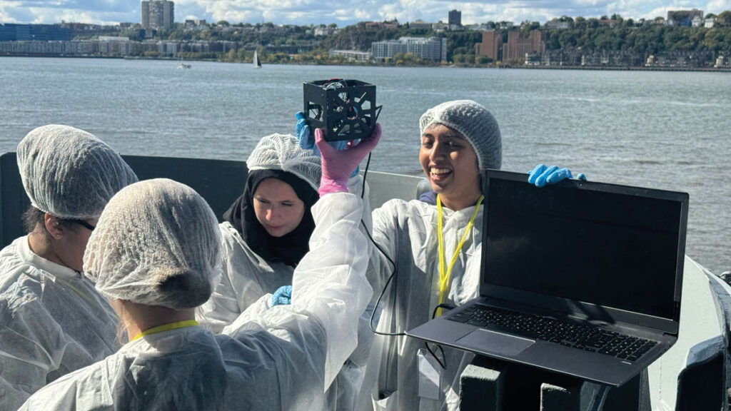 Two young people wearing protective clothing hold aloft a small cube connected to a laptop computer, while two other young people record data. They stand on the deck of a stationary ship on a river with city buildings and trees visible on the far riverbank.
