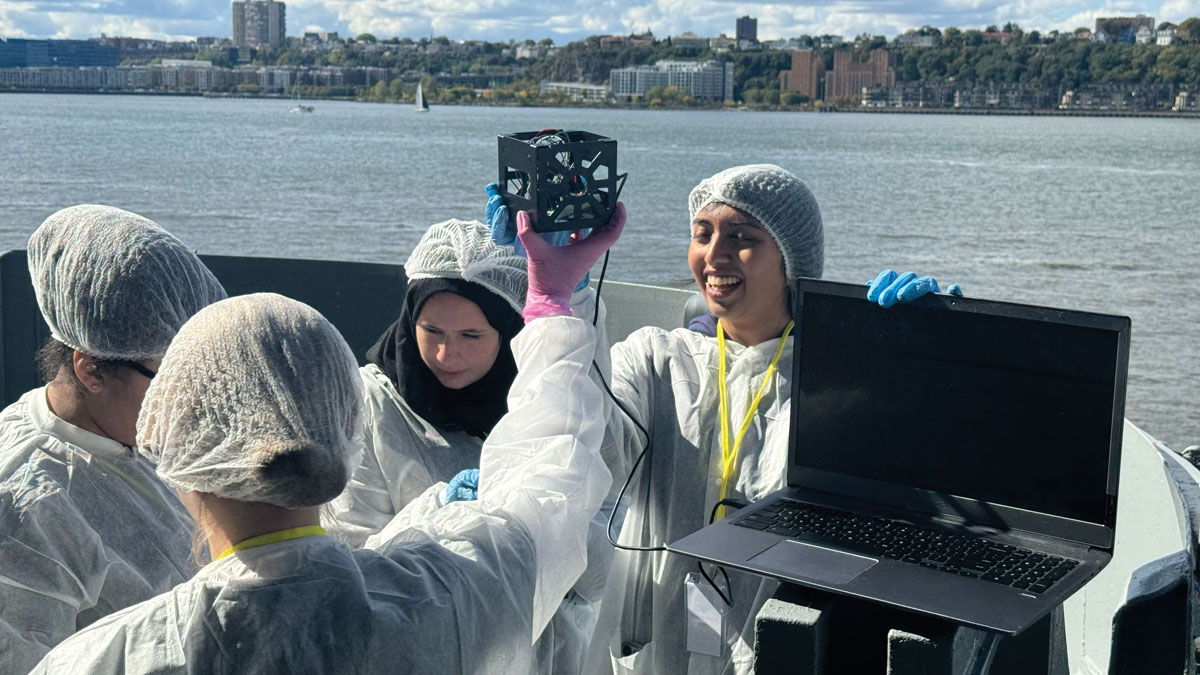 Two young people wearing protective clothing hold aloft a small cube connected to a laptop computer, while two other young people record data. They stand on the deck of a stationary ship on a river with city buildings and trees visible on the far riverbank.