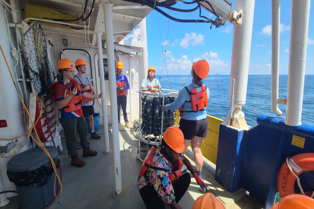 Six people wearing orange hard hats and life vests are on the deck of a ship.