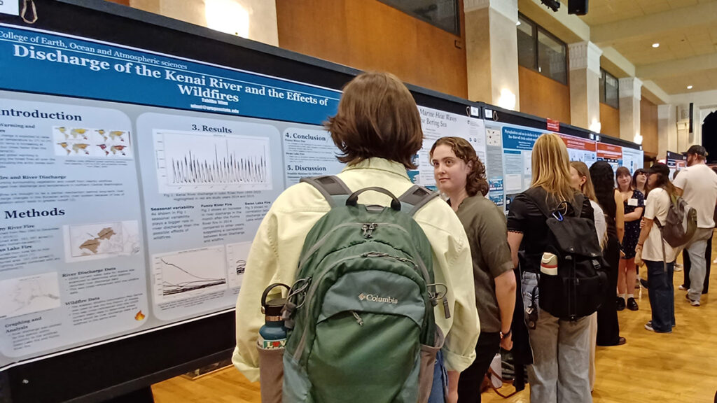 Students speak with one other in small groups while standing in an aisle between two lines of research posters.