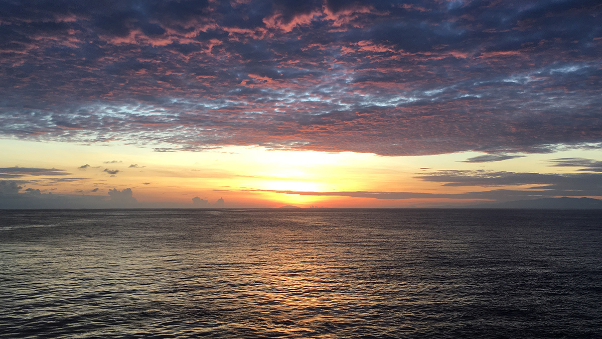 The Sun rises over the ocean. Purple clouds are in the sky above.