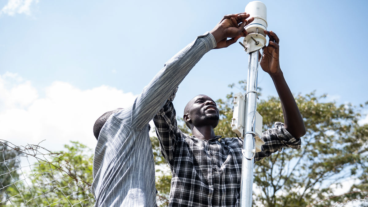 Two men install a weather station mounted on a tall metal pole.