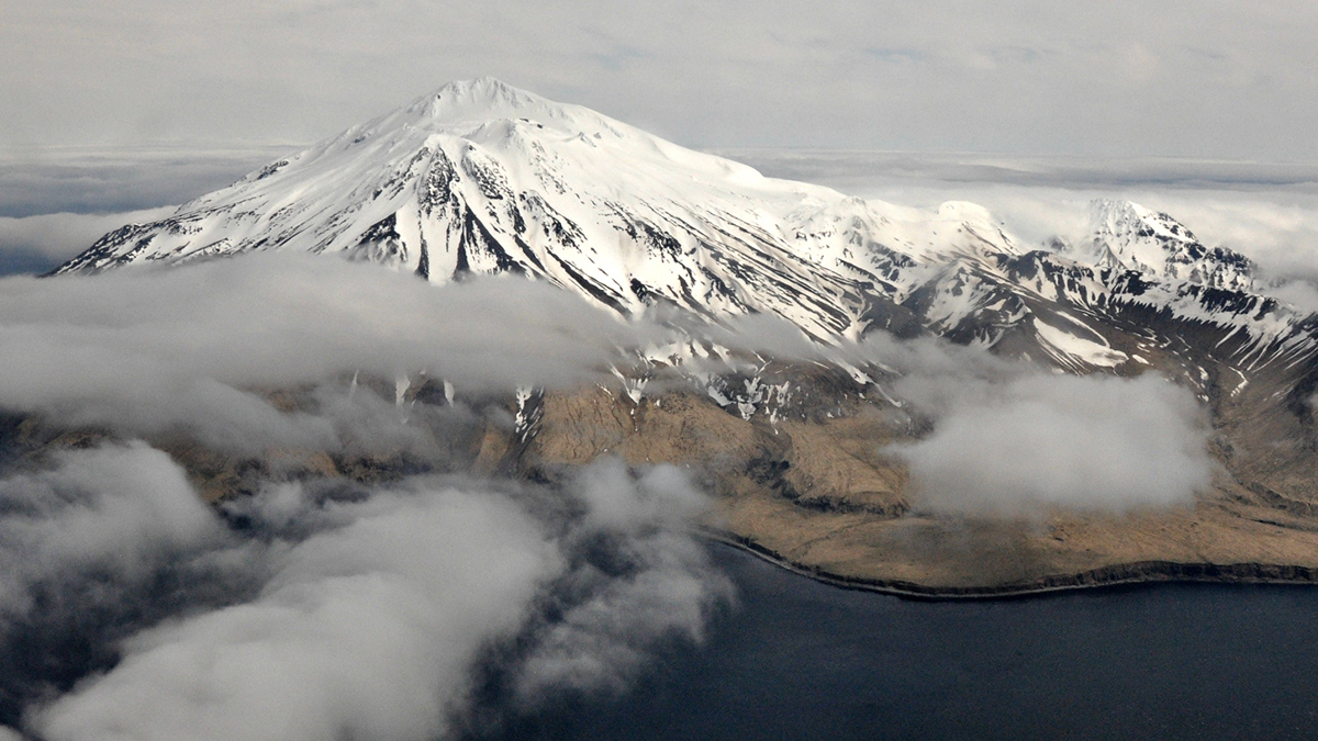 A snow-covered mountain on an island is seen from the air, through some parted clouds.