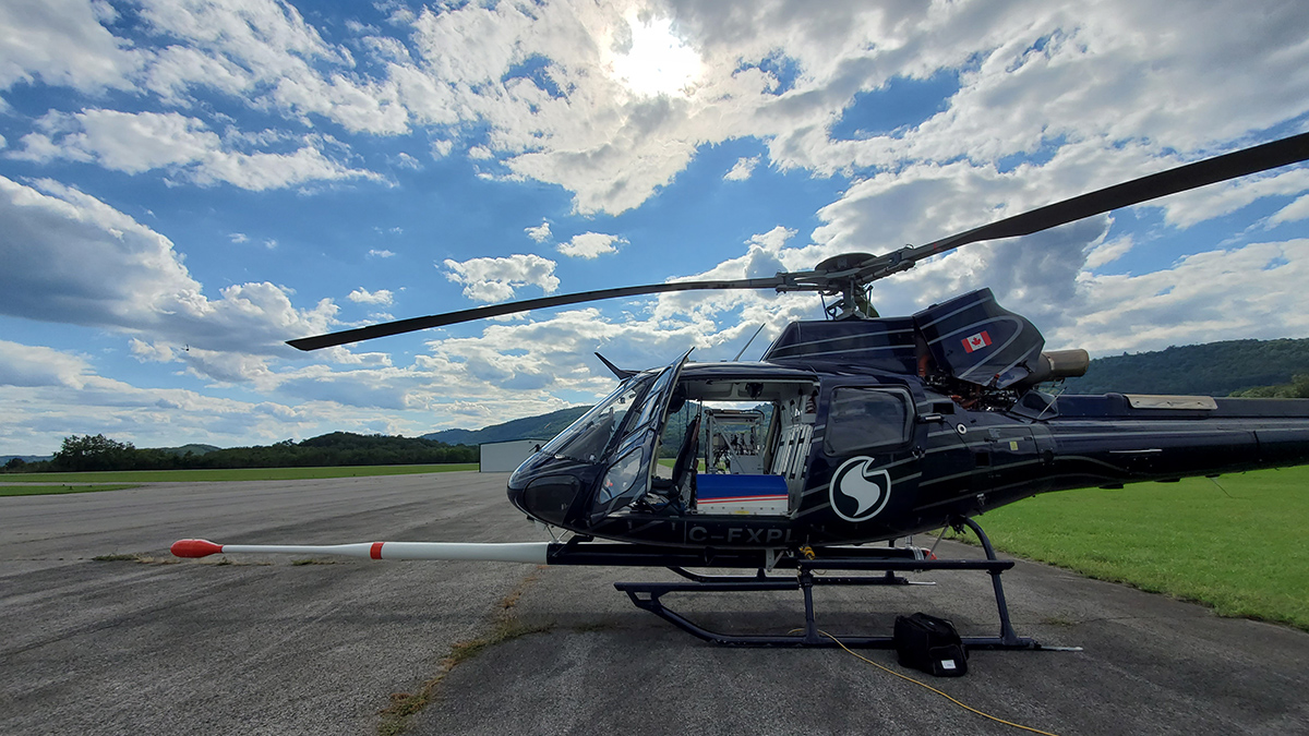 A helicopter equipped with a long boom-like sensor attached to the bottom and extending in front sits on asphalt tarmac under a partially cloudy blue sky.