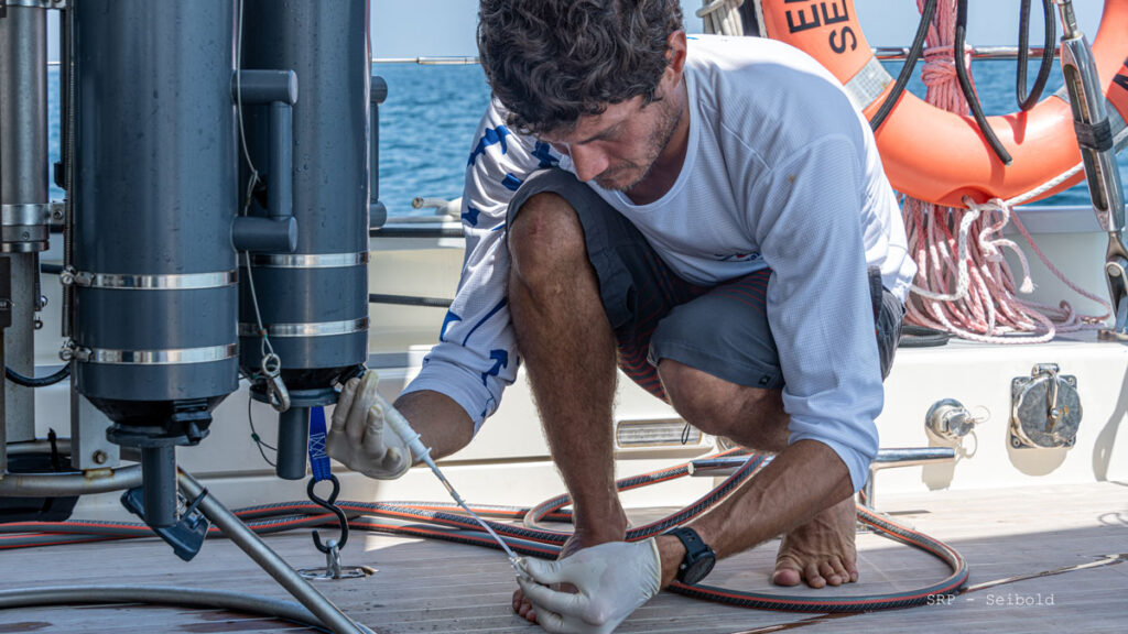 A man crouches on the deck of a ship. He is wearing latex gloves and collecting a water sample from a tank.