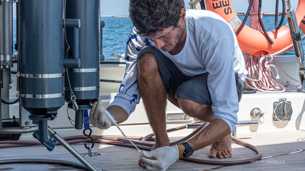 A man crouches on the deck of a ship. He is wearing latex gloves and collecting a water sample from a tank.