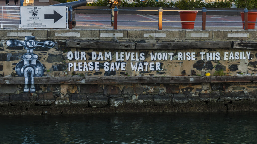 On the human-made wall of a waterway, appear graffiti of a water spigot beside the text “Our dam levels won’t rise this easily. Please save water.”