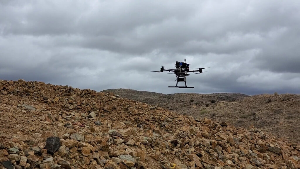 A helicopter-like drone hovers low over a large pile of rocky mining waste.
