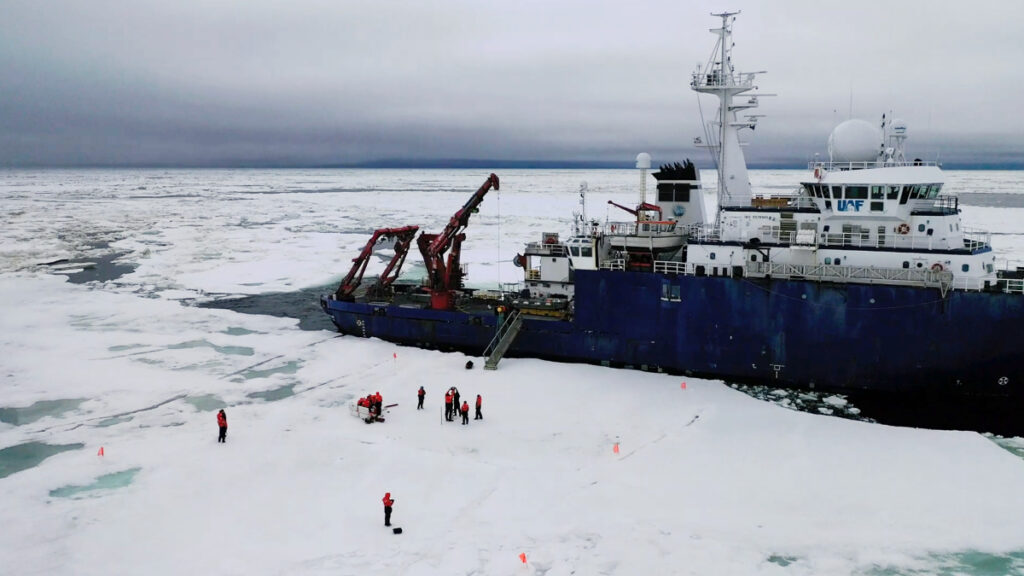 An aerial photo of a research team standing on ice with a research vessel looming in the background.