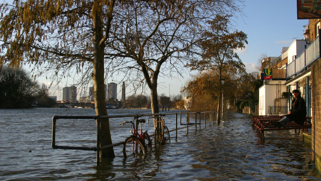 A man sits on the balcony of a flooded building along the banks of an overflowing river.