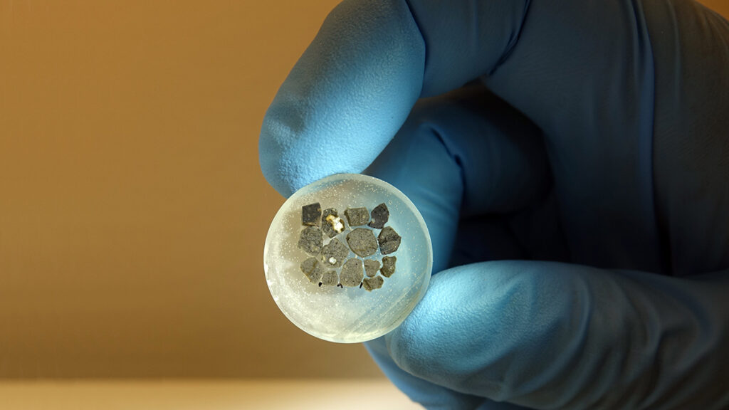 Pieces of gray rock are embedded in a small plastic disk and held by a hand wearing a blue glove.